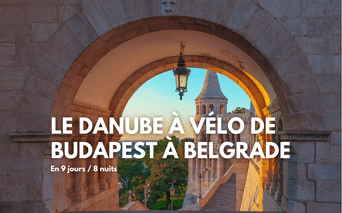 Vue à travers une arche en pierre avec lanterne suspendue, montrant un clocher et des arbres au coucher du soleil. Texte : Le Danube à vélo de Budapest à Belgrade.