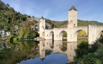 Pont médiéval Valentré avec trois tours et six arches enjambant le Lot, parfaitement reflété dans l'eau calme, entouré de collines vertes.