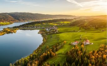 Vue aérienne du Lac Brenet entouré de forêts d'automne et de prairies vertes. Un petit village se niche le long du rivage avec des collines.