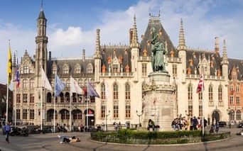 Palais Provincial de Bruges avec architecture gothique et flèches ornées. Un monument se dresse au centre de la place, entouré de touristes.