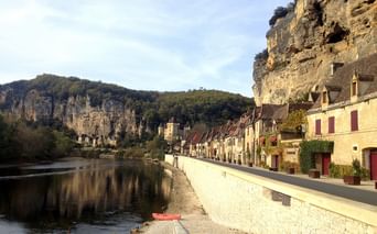 Maisons en pierre avec volets rouges le long d'une route au bord de la rivière en Périgord Noir, adossées à des falaises calcaires spectaculaires.