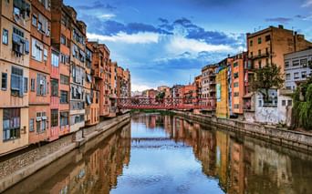 Des bâtiments colorés bordent les deux rives de la rivière Onyar à Gérone, avec un pont rouge traversant l'eau. Le ciel bleu avec des nuages blancs se reflète dans l'eau calme.