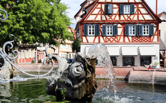 Fontaine ornée avec jet d'eau devant une maison à colombages rouge et blanc avec volets bleus dans une ville de la vallée du Rhin.