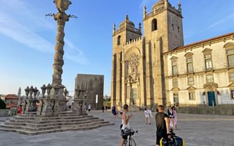 Cathédrale de Porto avec pilier en pierre orné Cathédrale de Porto avec tours jumelles et façade baroque, pilier en pierre orné au premier plan. Touristes et cyclistes sur la place.