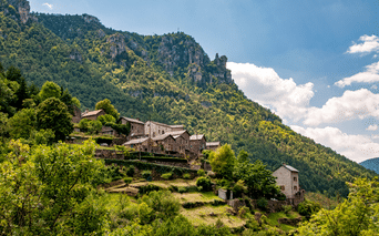 Village traditionnel en pierre sur une colline des Cévennes avec des champs en terrasses, entouré de forêts vertes et de pics rocheux.