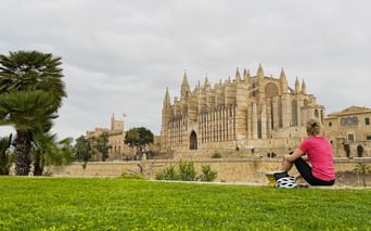 Cycliste en maillot rose assise sur l'herbe avec casque de vélo à côté, face à la cathédrale gothique de Palma à Majorque avec palmiers.