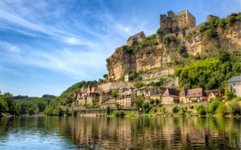 Village médiéval avec château en pierre perché sur falaise calcaire au-dessus de la Dordogne calme en Périgord. Maisons traditionnelles en cascade vers l'eau.