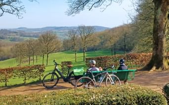 Deux cyclistes sur banc vert surplombant les collines des Flandres françaises. Deux vélos garés sous des arbres nus avec champs verts.