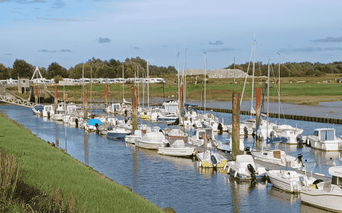 Petit port de plaisance avec voiliers blancs amarrés le long de quais en bois. Berges herbeuses encadrent le chenal, caravanes et dunes en fond.