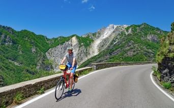 Cycliste avec casque et sacoches roulant sur route de montagne sinueuse avec garde-fou en pierre, entouré de montagnes boisées sous ciel bleu.