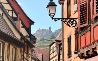 Maisons à colombages traditionnelles sur la Route des Vins d'Alsace avec lampadaire orné et château sur colline avec vignobles en arrière-plan.