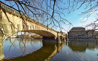 Pont en pierre avec arches enjambant la Vltava à Prague, avec le théâtre national visible sur la rive droite sous des arbres nus d'hiver.