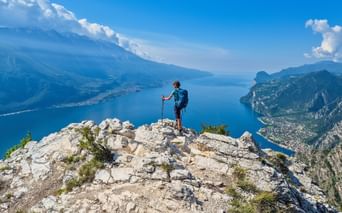 Randonneur avec sac à dos et bâtons sur sommet rocheux de Punta Larici, vue sur eaux bleues du lac de Garde entouré de montagnes.
