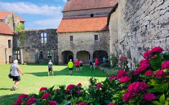 Cyclistes explorant une cour médiévale en pierre en Franche-Comté avec des passages voûtés et des fleurs roses au premier plan sous un ciel bleu.