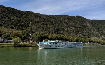 Bateau de croisière MS Switzerland naviguant sur eau verte calme avec collines boisées et petit village en arrière-plan sous ciel bleu.