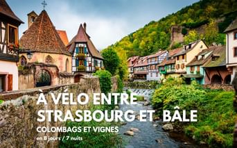 Maisons à colombages colorées bordent un ruisseau dans un village alsacien. Une chapelle au toit pointu se dresse à gauche, collines boisées derrière.