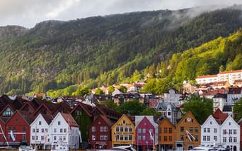 Rangée de maisons en bois colorées traditionnelles au bord de l'eau à Bergen, Norvège, avec montagnes boisées et nuages bas en arrière-plan.