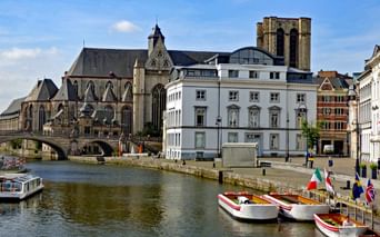 Front de mer historique de Gand montrant une cathédrale gothique avec haute tour, des bâtiments classiques blancs et des bateaux touristiques amarrés le long du canal.