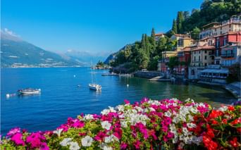 Fleurs roses, blanches et rouges encadrant le lac de Côme avec bateaux sur l'eau bleue, bâtiments colorés sur la colline et montagnes en arrière-plan.