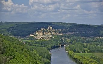 Château de Beynac perché sur une colline surplombant la Dordogne. Un pont de pierre traverse la rivière, entouré de forêts et collines vertes.