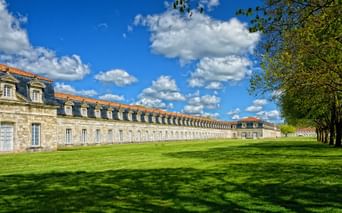 Long bâtiment historique en pierre Corderie Royale avec lucarnes et toit de tuiles orangées, devant pelouse verte et allée bordée d'arbres sous ciel bleu.