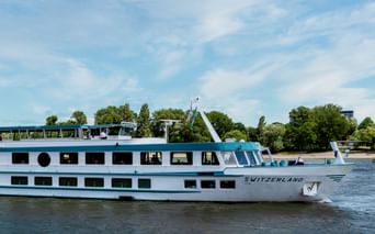 Bateau de croisière fluviale blanc MS Switzerland naviguant sur eau calme avec deux ponts de fenêtres, arbres verts et ciel bleu en arrière-plan.