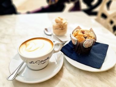 Cappuccino avec latte art dans une tasse de marque sur soucoupe blanche, à côté d'un muffin aux pépites de chocolat sur serviette bleue.