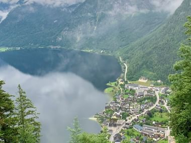 Vue aérienne du village de Hallstatt niché au bord d'un lac alpin, entouré de montagnes brumeuses. Rails de funiculaire visibles au premier plan.