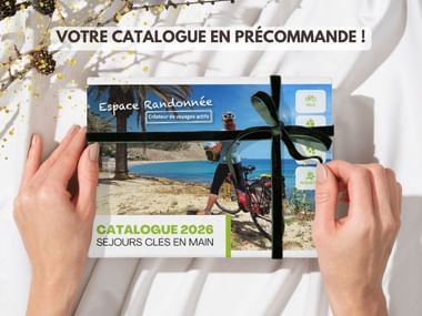 Mains tenant le catalogue Espace Randonnée 2026 avec ruban noir, montrant un cycliste sur la plage. Texte 'Votre catalogue en précommande'.
