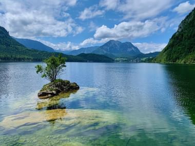 Lac cristallin d'Altaussee avec petite île rocheuse et arbre solitaire, entouré de montagnes boisées sous un ciel nuageux.