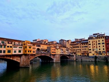 Pont Ponte Vecchio à Florence avec bâtiments colorés enjambant l'Arno. Arches en pierre reflétées dans l'eau calme sous le ciel du soir.