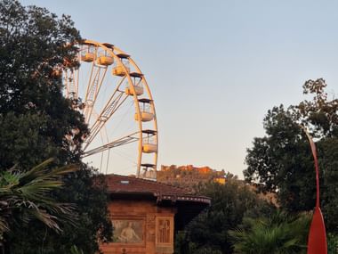 Grande roue jaune s'élevant au-dessus des arbres et d'un bâtiment à Montecatini, Toscane, avec une forteresse sur une colline visible en arrière-plan au coucher du soleil.