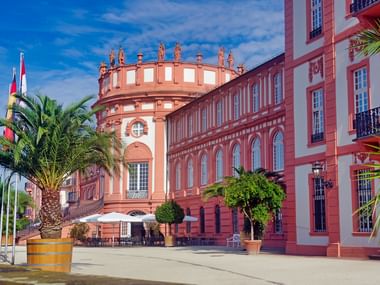 Château baroque rose à Eltville avec tour ronde distinctive surmontée de créneaux décoratifs. Palmiers et drapeaux encadrent l'entrée de la cour.
