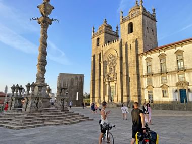 Cathédrale de Porto avec tours jumelles et façade baroque, pilier en pierre orné au premier plan. Touristes et cyclistes sur la place.