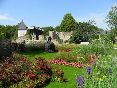 Jardin paysager à Coblence avec parterres de fleurs colorés, pelouse verte et sculpture sombre. Ruines en pierre et tour blanche en arrière-plan.