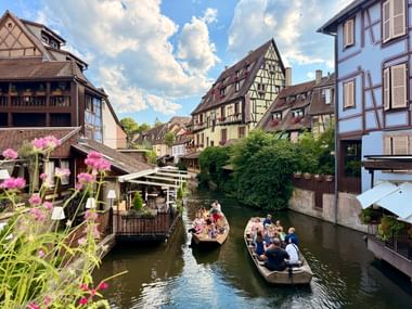 Deux bateaux avec touristes sur un canal entre maisons à colombages traditionnelles sur la route des vins d'Alsace. Des fleurs roses encadrent la scène.