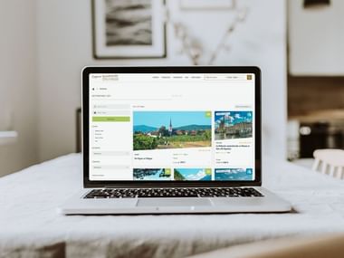 Ordinateur portable sur bureau blanc affichant site de randonnées avec images de vignobles et villes européennes dans salon lumineux.