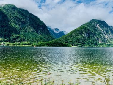 Lac de montagne clair entouré de collines boisées dans la région du Salzkammergut. La végétation verte pousse le long du rivage sous un ciel nuageux.