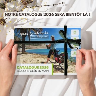 Mains tenant un catalogue cycliste 2026 avec ruban noir, montrant une scène de plage avec vélo. Texte : 'Notre catalogue 2026 sera bientôt là'.