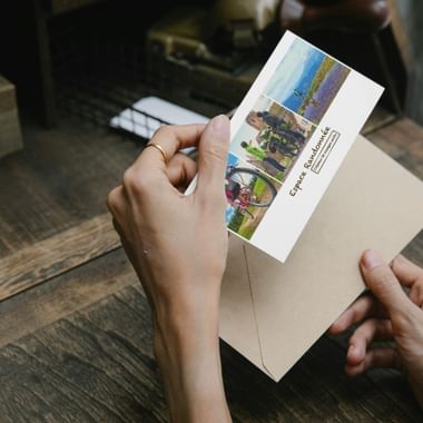 Mains tenant un bon cadeau avec trois photos de randonnée montrant des paysages de montagne et des randonneurs, posé sur une enveloppe beige sur une table en bois.