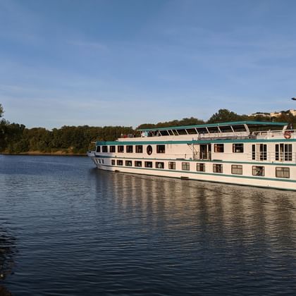 Bateau de croisière MS Switzerland naviguant sur un fleuve calme avec des arbres verts sur la rive gauche et des bâtiments visibles au loin.
