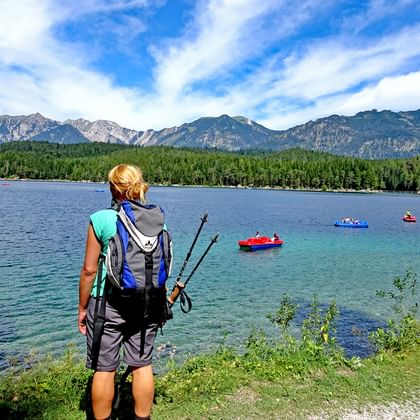 Randonneuse avec sac à dos et bâtons au bord du lac Eibsee, contemplant le lac turquoise avec pédalos et montagnes alpines.