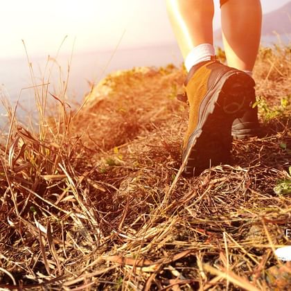 Gros plan sur les jambes et les chaussures de randonnée d'une marcheuse qui s'avance dans un paysage maritime sous un soleil brûlant