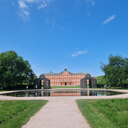 Château de Rastatt avec bassin Château de Rastatt, bâtiment baroque rouge avec dôme central, se reflète dans un bassin rectangulaire. Pelouses vertes et arbres matures encadrent la scène sous ciel bleu.