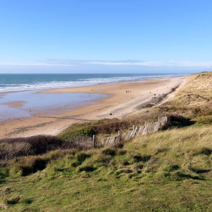 Large plage de sable à Hatainville avec dunes herbeuses, clôtures en bois et mer bleue calme sous ciel dégagé dans le Cotentin, France.