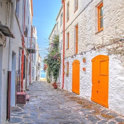 Ruelle pavée étroite à Cadaqués avec bâtiments blanchis à la chaux et portes et fenêtres orange vif, menant vers la mer sous ciel bleu.