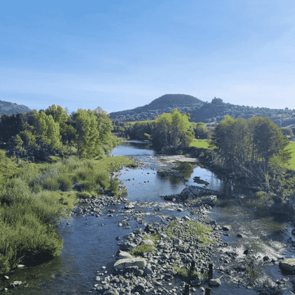 Rivière rocailleuse traversant un paysage verdoyant avec des arbres sur les deux rives. Collines et montagnes lointaines sous ciel bleu.