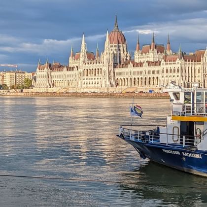 Bateau de croisière MS Princess Katharina sur le Danube avec le Parlement hongrois en arrière-plan sous un ciel nuageux.