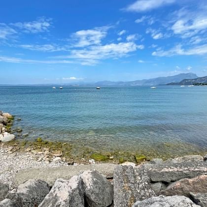 Rivage rocheux du lac de Garde près de Bardolino avec eau bleue claire, brise-lames en pierre et montagnes en arrière-plan sous ciel nuageux.
