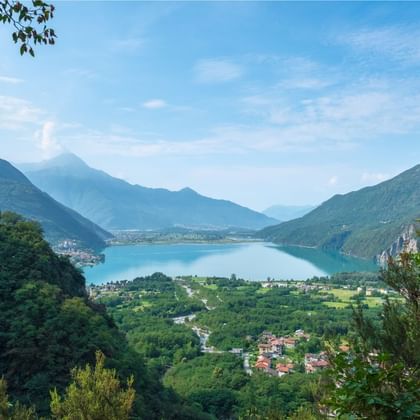 Lac de Mezzola niché dans une vallée entre des montagnes vertes. Un village avec des maisons au premier plan, entouré de végétation luxuriante.